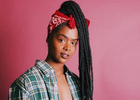 Elegantly posed young woman with braided hair and plaid shirt against a pink background.